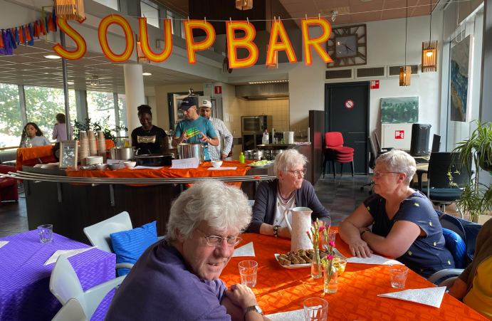 Buurtbewoners aan een ronde tafel met op de achtergrond personeel achter de soupbar.